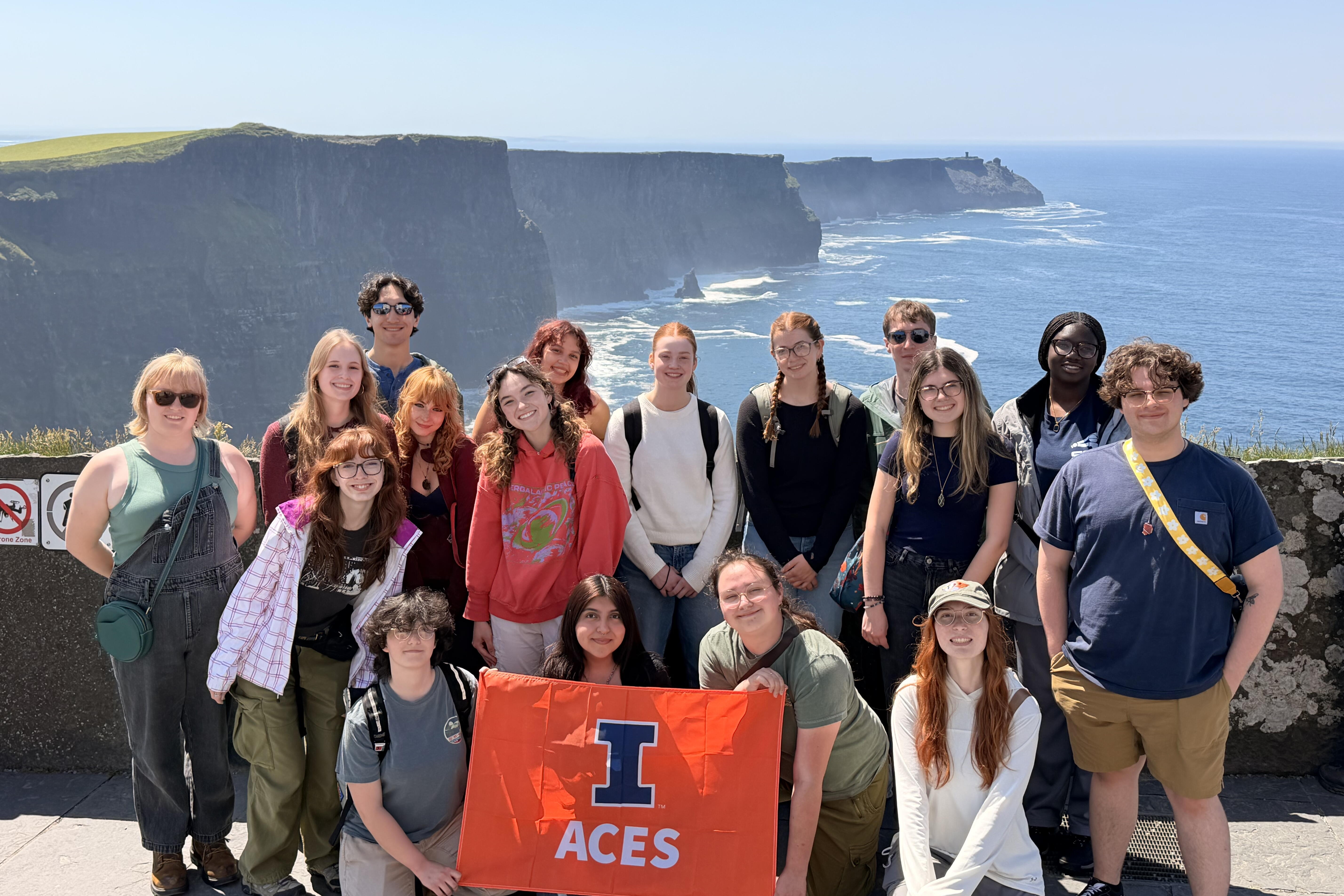 A group of students in the foreground hold an orange banner with the ACES logo. Behind them, the ocean appears below large cliffs.