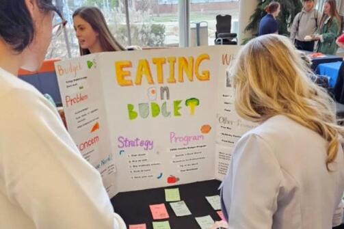 two people standing in front of a table with a standing cardboard poster that reads eating on budget