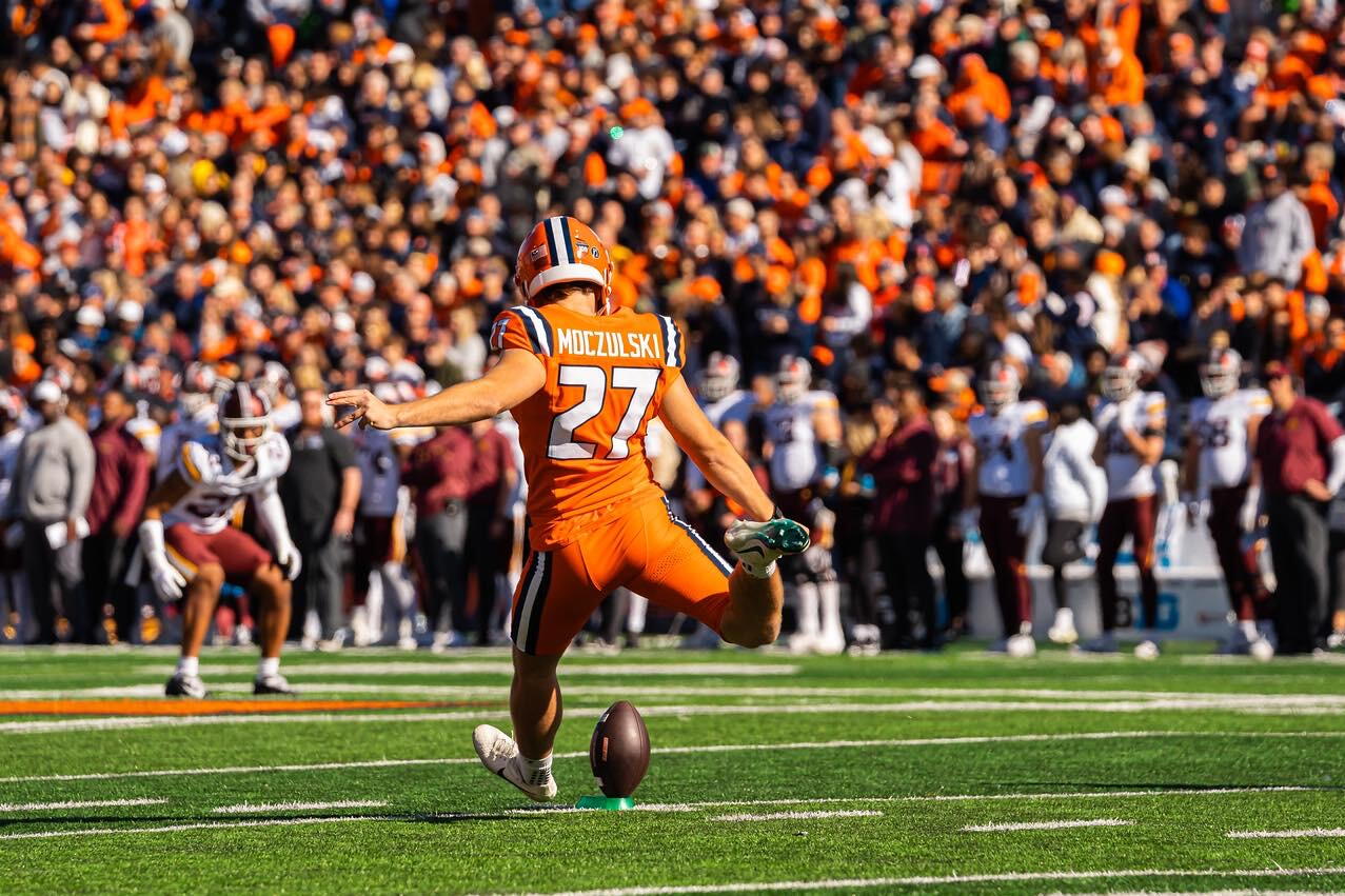 Ethan Moczulski kicking a football before a sea of fans in orange.