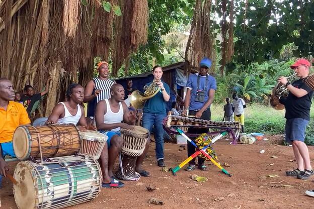 A diverse group of people playing various musical instruments, including drums, xylophone, and horns, under a large tree in an outdoor setting.