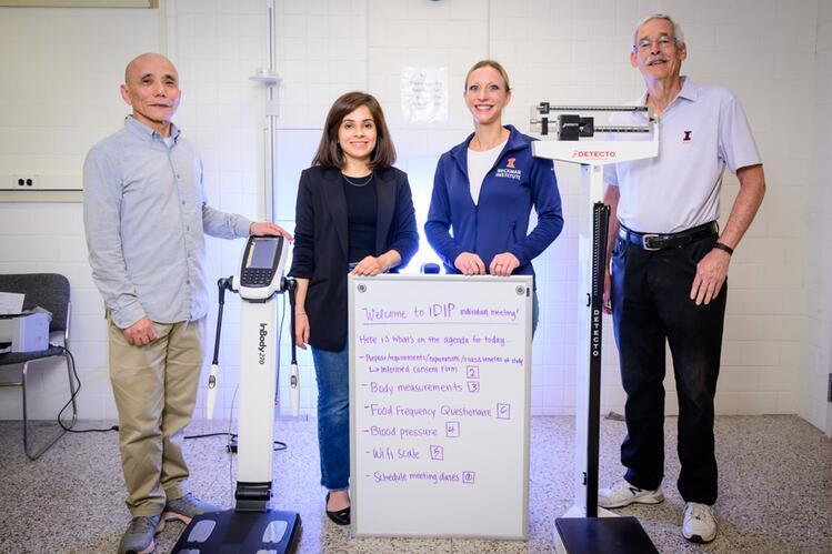 Four people standing around scales and a white board with text  