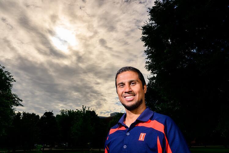 A man in a blue and orange shirt standing outside with a background of trees and clouds