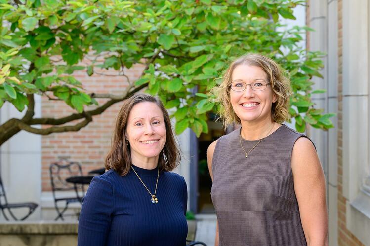 Two women smiling at the camera, standing outdoors in front of a building and a tree