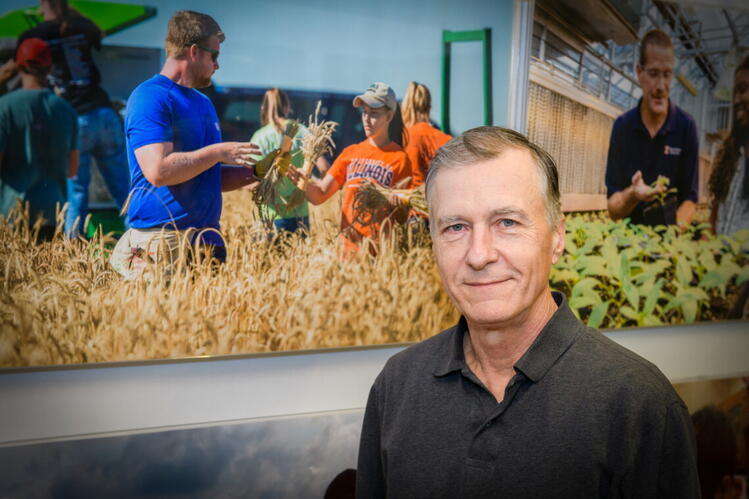 Lowell Gentry stands before photos of people standing in golden brown crop fields and in a greenhouse