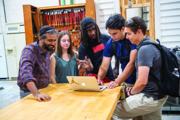 Four students and a professor discuss work around a laptop