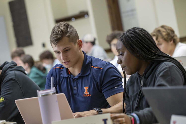 Two students are looking at a laptop computer in a classroom with other students visible in the background