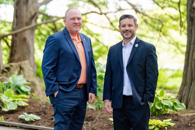 Two men in suits stand outdoors. Plants and trees are in the background.