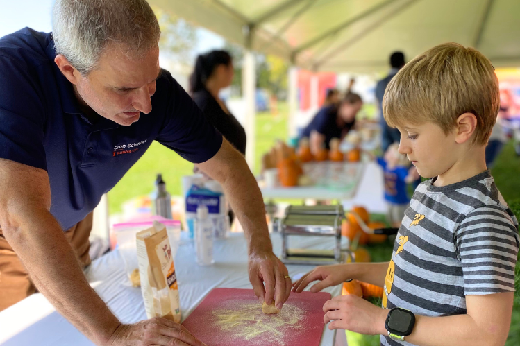 An adult assists a young boy in making pasta at a table under a tent during an outdoor event. Cooking supplies and ingredients are visible on the table.