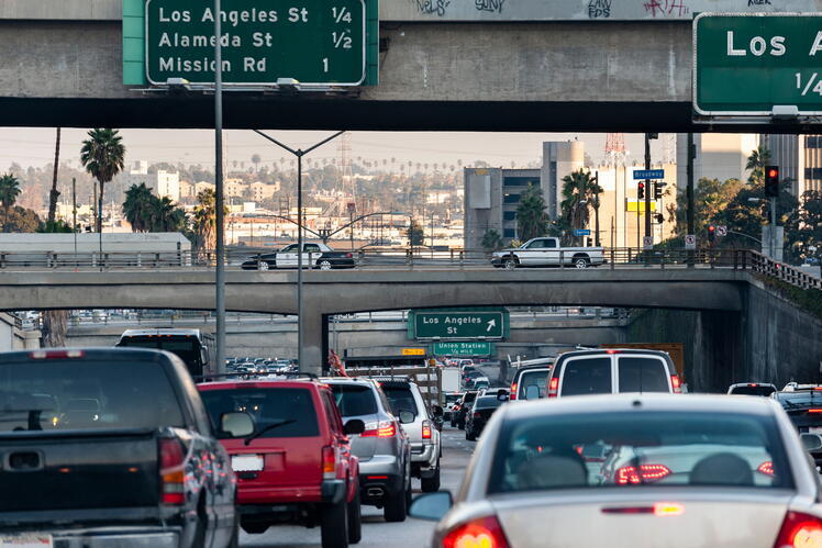 Lines of cars stopped on a Los Angeles freeway. Local road signs are visible on bridges above the cars, and palm trees and buildings are in the background.