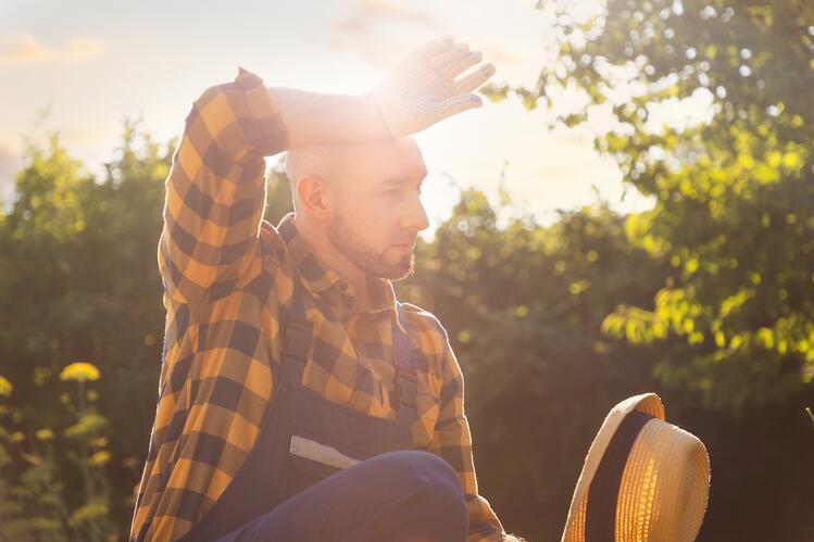 A person in a plaid shirt and overalls shields their eyes from the sun with a gloved hand while holding a hat. Lush greenery surrounds them.