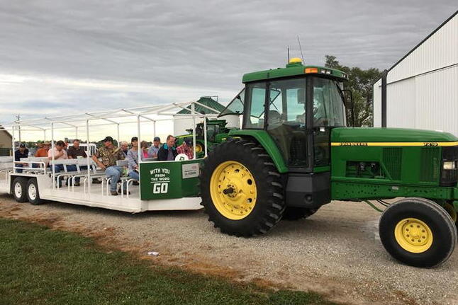 A green tractor pulls a covered wagon with seated passengers on a gravel path near a white building under a cloudy sky