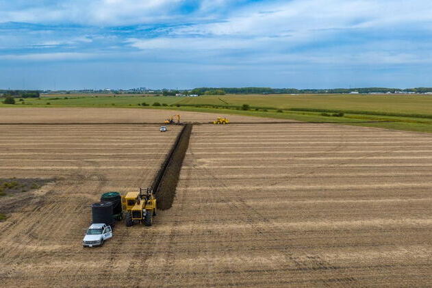 An aerial view of farm fields. In the foreground, a yellow tractor and white car are parked next to two large reels of black drainage pipe