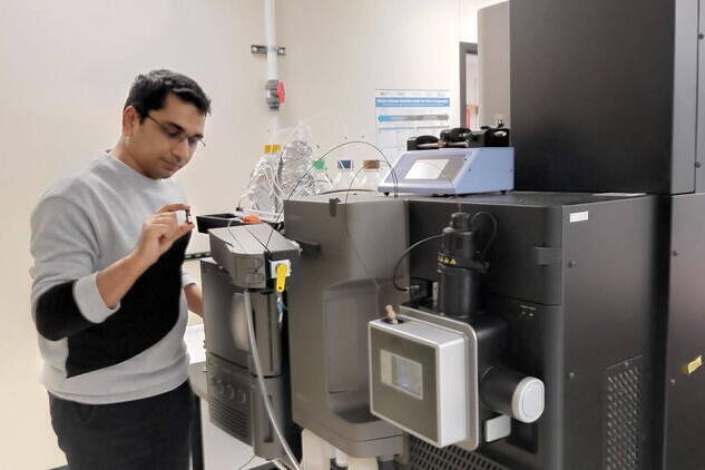 a man wearing glasses stands next to a large piece of laboratory equipment, holding a sample.
