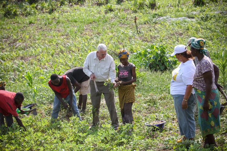 Burt Swanson speaks to a woman holding a plant, while both stand in a green field