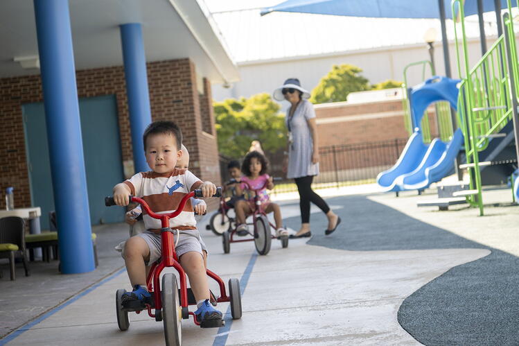 A child riding a tricycle in a playground space with other children and a teacher in the background