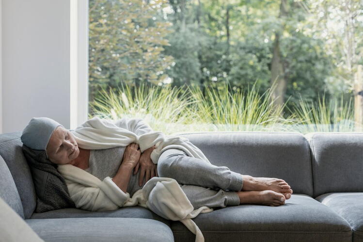A woman wearing a head scarf, robe, and pajamas lies curled up on a couch, clutching her stomach. In the background, trees and grasses are visible through a window.