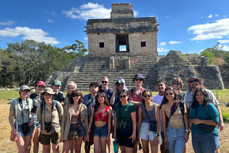 A group of young adults wearing sunglasses, hats, and t-shirts pose in front of a Mayan ruin in the Yucatán Peninsula.