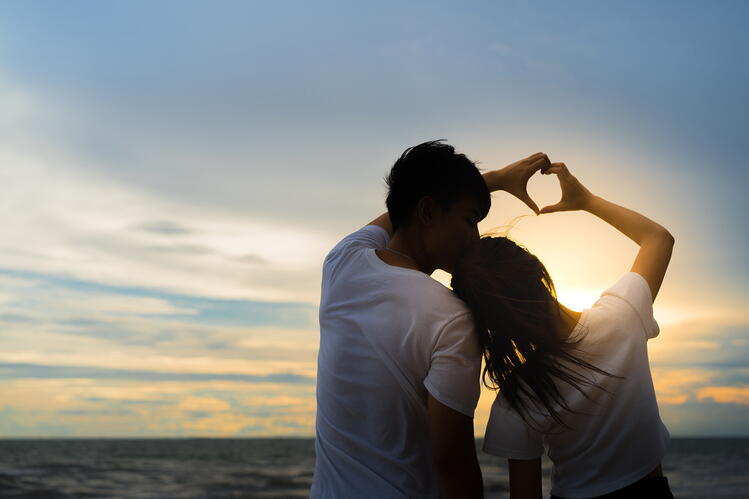 A couple standing by a sunsetm, their backs to the camera, holding hands in the shape of a heart