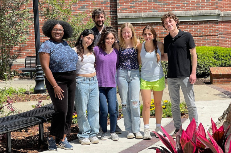Terence Crofts’ research group, including paper co-authors Ezabelle Frank (far left), Terence Crofts (behind the group), Elizabeth Bernate (center, in purple), and Hayden Allman (far right). Six young adults stand outdoors with a brick building in the background. A bearded man stands just behind the group.