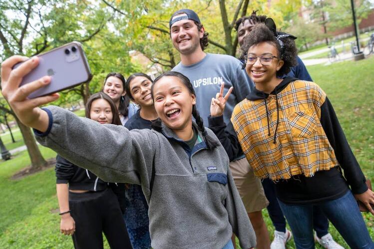 A group of students pose together for a selfie. A green lawn and trees are in the background.