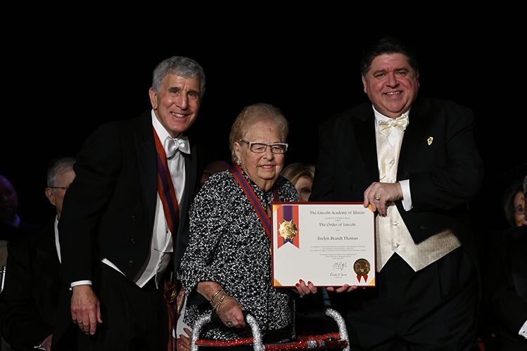 Evelyn Brandt Thomas with a walker receives an award certificate on stage, flanked by two men in formal attire holding the document.