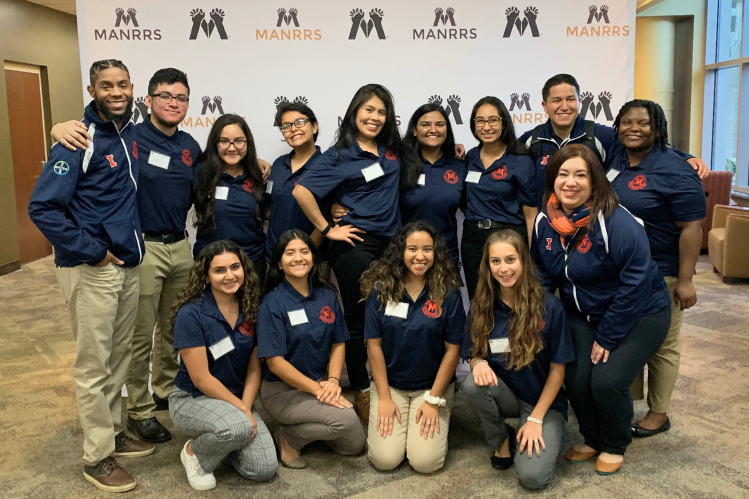 Group of people in matching blue and orange shirts posing together in front of a MANRRS backdrop.