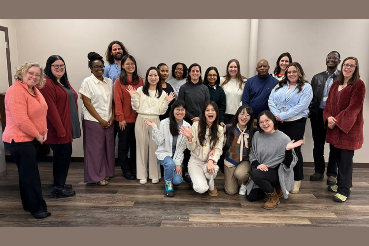 A group of 20 people pose together indoors, standing and smiling at the camera in a room with light walls and wood flooring.