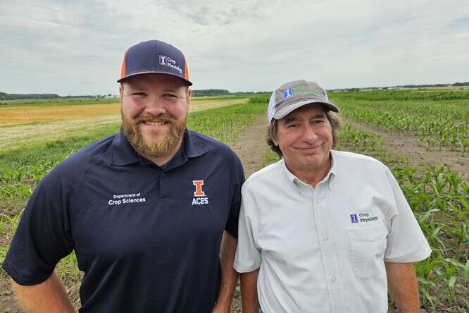 Two men in branded shirts and hats stand in front of a cornfield, smiling. The field has young corn plants under a partly cloudy sky.