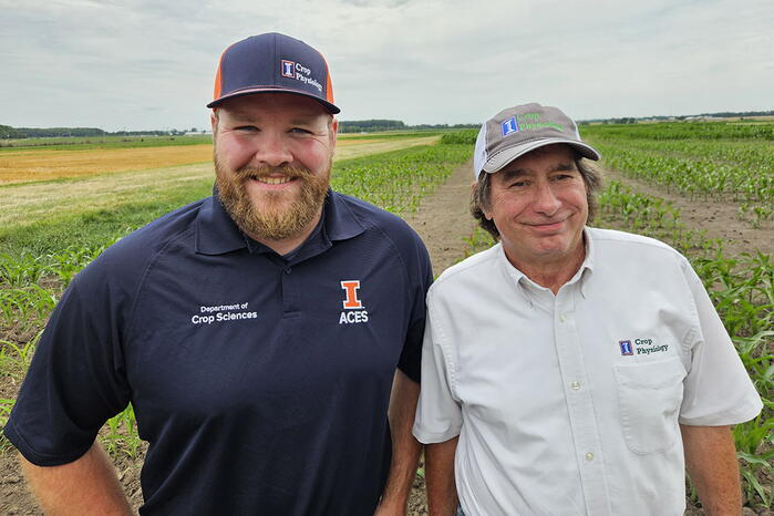 Two men in hats and university-branded t-shirts stand in the foreground. The background is a crop field.