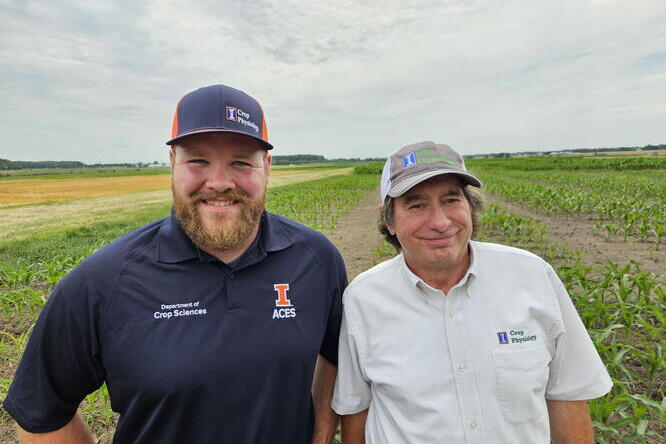 Two men smile in front of a field of crops.