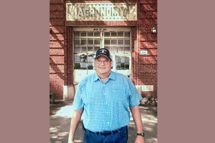 Man in blue shirt and baseball cap stands in front of an old brick building with the word Agronomy over the door