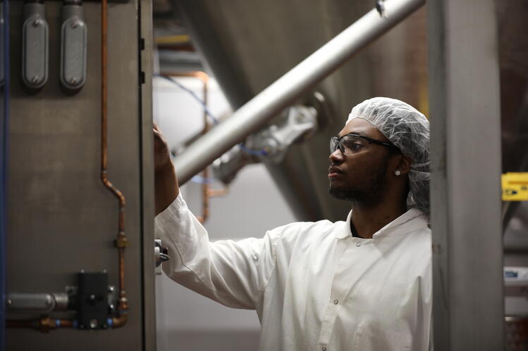 A man in a white lab coat and hairnet stands between metal machines in the Pilot Plant.