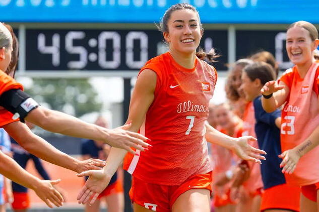 A female Illinois soccer player in an orange uniform high-fives teammates on the field, with a scoreboard in the background showing 45:00.