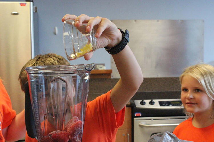 A boy is pouring fruit into a blender while a girl is looking on. They are sitting in a kitchen with a stove in the background.