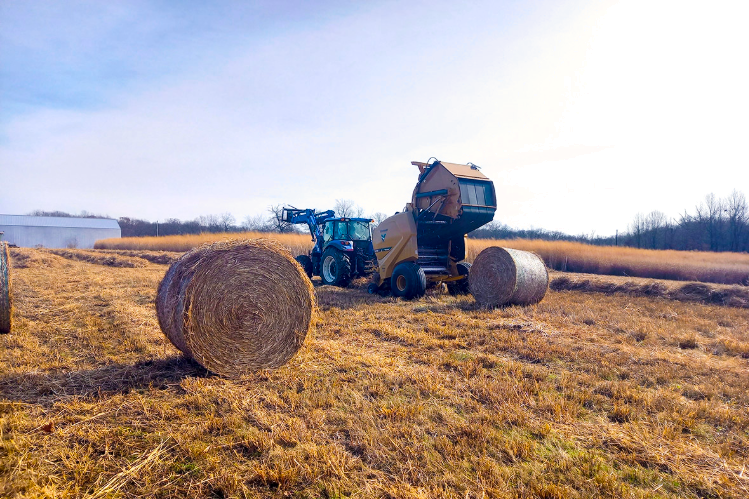 A tractor operates a round baler in a field, producing large cylindrical hay bales on a sunny day.