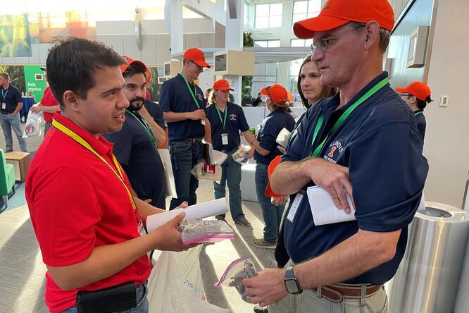 Two men are talking while holding documents and bags of items. One is wearing a red shirt, the other a blue shirt and orange cap. Other people in similar attire are visible in the background.