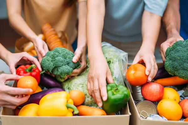 Three people with only their arms and hands visible are handling fresh, colorful vegetables in boxes