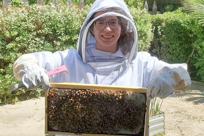 A woman in a white beekeeper outfit is holding a frame of bees, standing in an outdoor setting with trees in the background. 