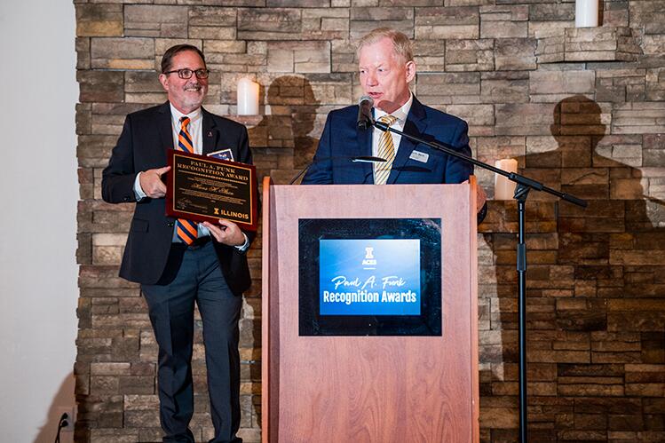 Hans Stein speaks at the podium with dean Germán Bollero in the background holding the plaque.