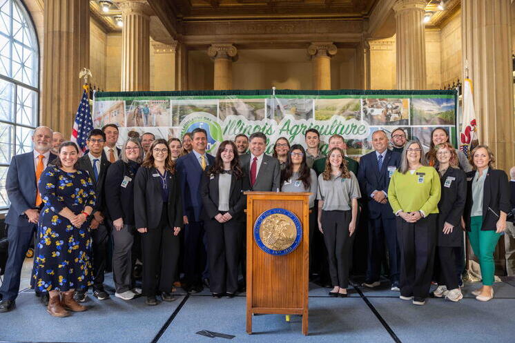 Faculty and students stand with Illinois legislative members on stage in front of a large banner showcasing Illinois agriculture.