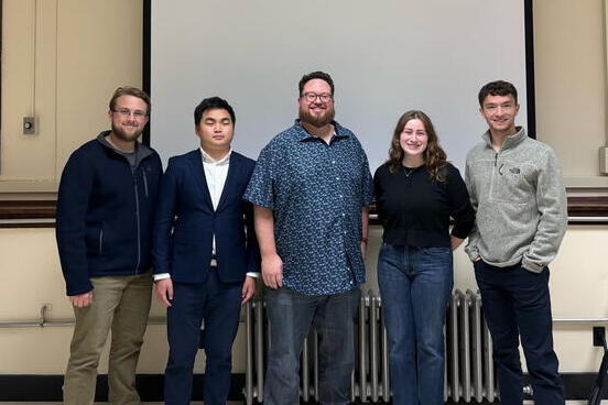 Five individuals stand in front of a screen and pose for a photo