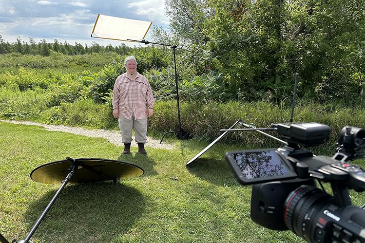 A woman in a checked shirt and boots lectures on camera outdoors.