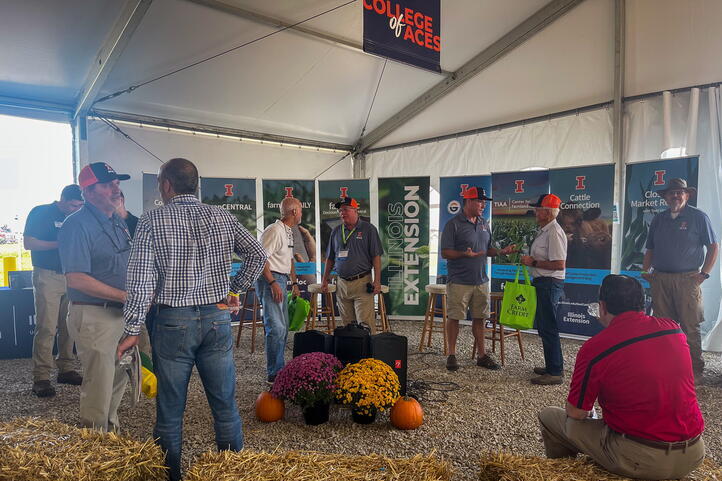 A large group pictured inside the tent chatting in pairs surrounded by U. of I. and ACES branding.