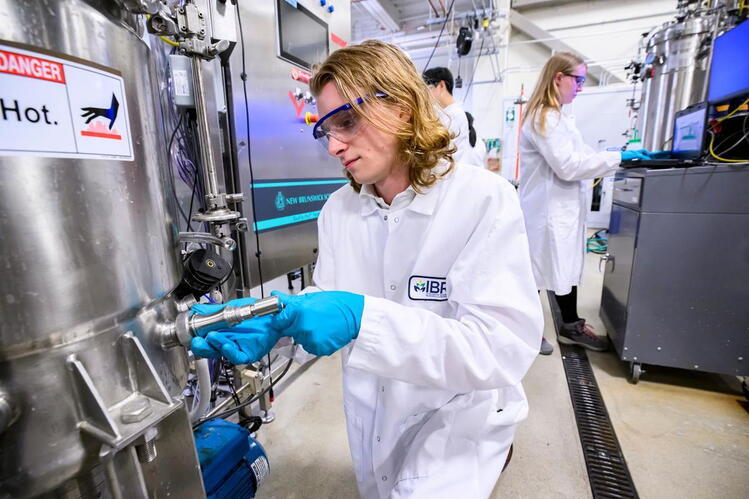 A person wearing safety goggles and gloves adjusts a valve on industrial equipment in a laboratory, with others working in the background.