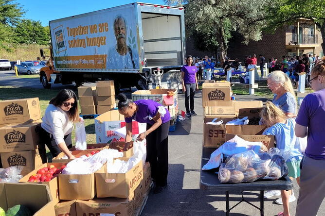 Volunteers distribute fresh produce and groceries from large boxes outdoors, with a truck labeled "Together we are solving hunger" in the background.