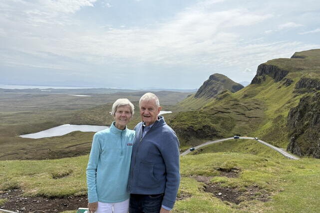 A woman in a light blue shirt and a man in a dark jacket stand in front of a grassy, mountainous landscape