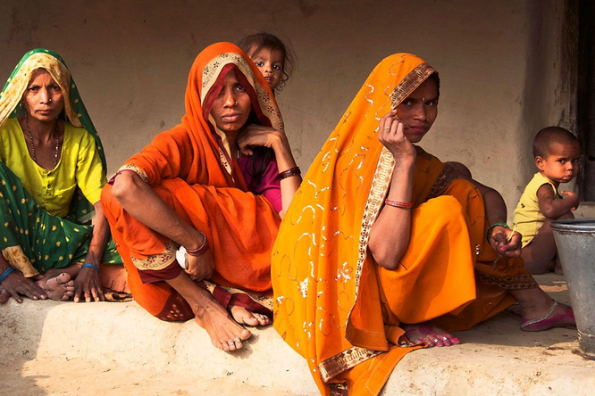 A group of women and children in brightly colored, traditional clothing sitting on a stone floor outside a building