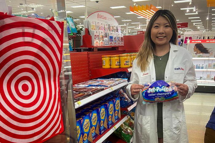 Woman in lab coat stands in Target checkout aisle holding a pack of marshmallows.