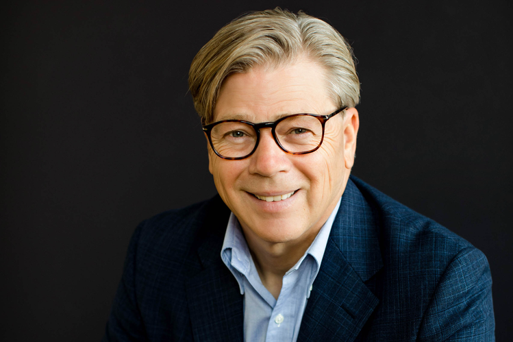 Portrait of a smiling Doyle Karr with glasses, wearing a blue shirt and dark blazer, against a black background.