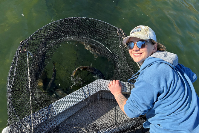 A woman in a baseball hat and hoodie leans over the side of a boat with a large net structure containing dark-colored fish. Lake water fills the background of the image.